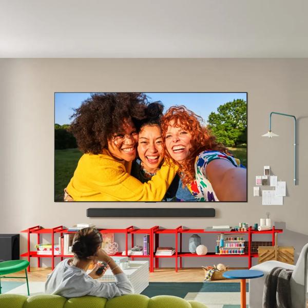 An LG TV and soundbar appear in a colorful living room. A vivid image of 3 women appears on the screen as a man watches from his couch. 