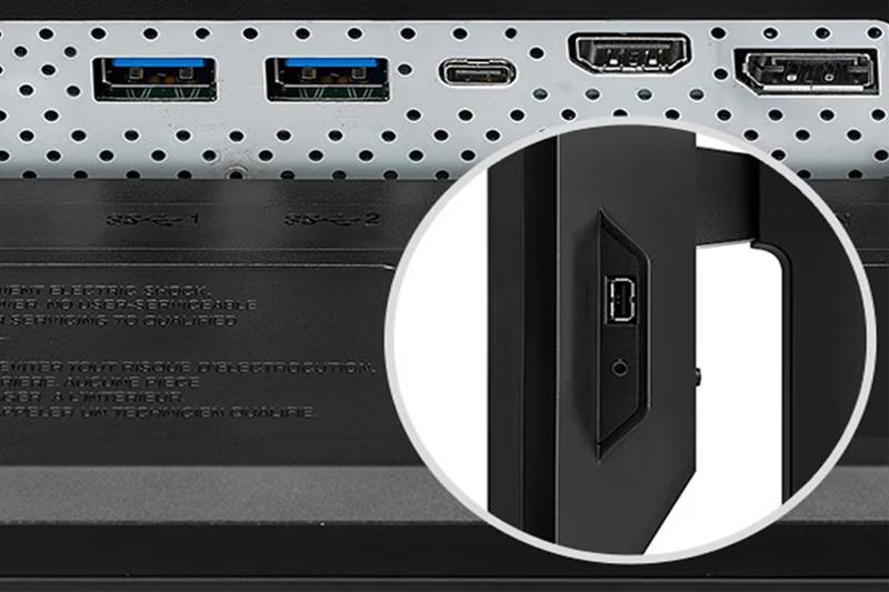 The image shows a detailed view of a computer monitor's connectivity ports. At the top section, there are two USB 3.0 ports, a USB-C port, and two HDMI ports arranged horizontally on a perforated metal plate. Below the ports are small embossed symbols for each USB port labeled "1" and "2." The background is textured with tiny holes, suggesting ventilation. The lower part shows warning text with an inset circle zooming in on the side of the monitor. This inset highlights an additional USB port and an audio jack on the side panel.