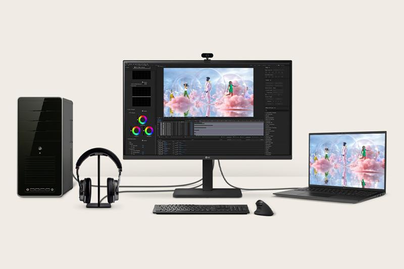 The image shows a computer workstation setup on a light beige background. At the center is a large flat-screen monitor displaying a colorful image editing interface with three individuals standing among clouds and reflections. To the left of the monitor is a black desktop computer tower with a glossy front panel. A pair of over-ear headphones rests on a stand between the computer tower and the monitor. A keyboard and mouse are placed directly in front of the monitor, on the desk surface. To the right of the central monitor, a slim laptop displays the same image on its screen, showing a seamless extension of the digital workspace. Cables are visibly connecting the devices.
