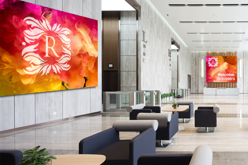 An upscale modern hotel lobby with two large LG DVLED Ultimate Business Displays. One is on the wall, and one is above the check-in counter. Both show bright orange, red, and yellow swirls with a white hotel logo overlaid on them. The one over the counter also says Welcome, Travelers.
