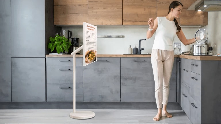 Woman looks at recipe on StanbyMe 2 touchscreen while preparing dinner in bright kitchen setting. Pop-out image of StanbyME 2 on stand and in folded mode.