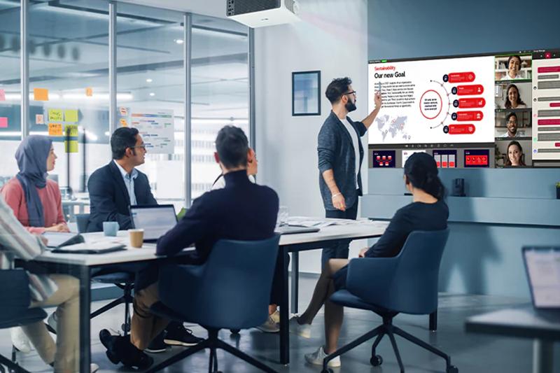 A group of professionals in a modern sleek meeting room. An LG ProBeam projector is suspended from the ceiling, projecting a presentation slide onto the wall of the meeting room. 