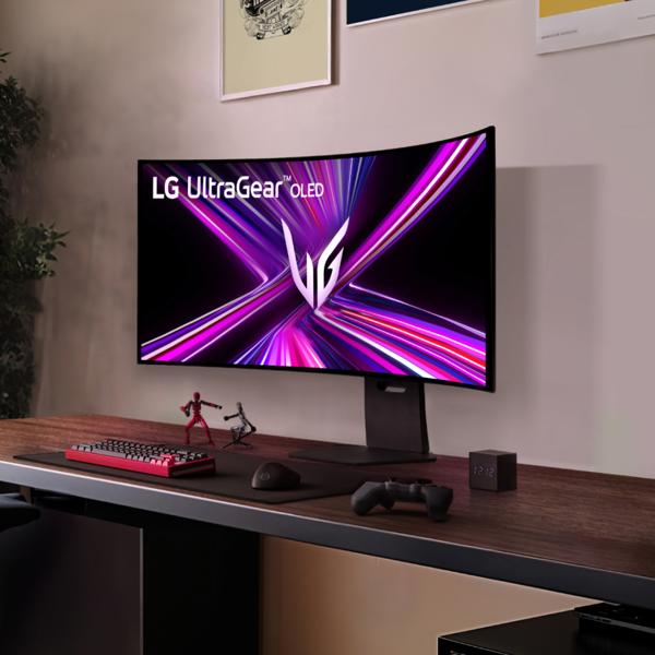 The LG 45 Inch UltraGear™ OLED Monitor on a desk with the UltraGear logo on the screen and a keyboard, action figures and a gaming controller in front of it.