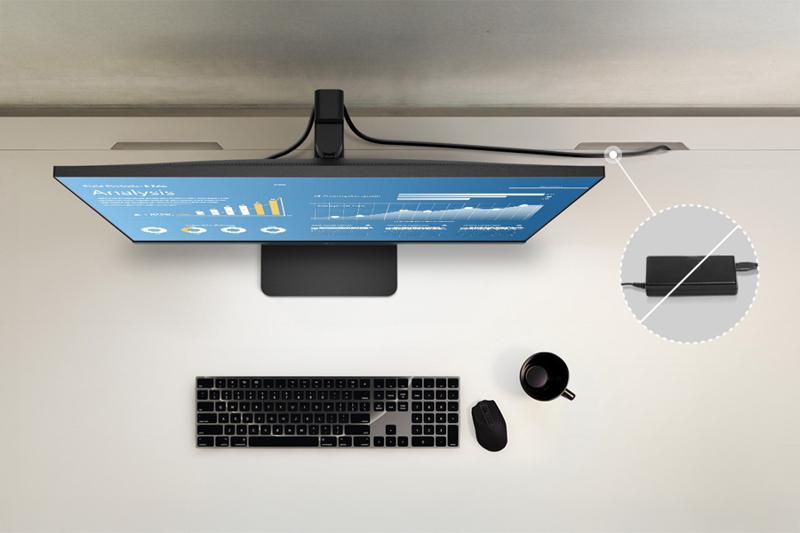 The image depicts an overhead view of a minimalistic workspace. At the center is a widescreen monitor displaying various graphs and charts on a blue background, suggesting data analysis or presentations. Below the monitor, there is a black keyboard and a wireless mouse placed on a light-colored desk. To the right of the mouse, a black cup is positioned, possibly for coffee or tea. A cable extends from the monitor towards the back, highlighted by a close-up inset showing a focused view of a cable connection.