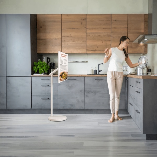 Woman looks at recipe on StanbyMe 2 touchscreen while preparing dinner in bright kitchen setting. Pop-out image of StanbyME 2 on stand and in folded mode.