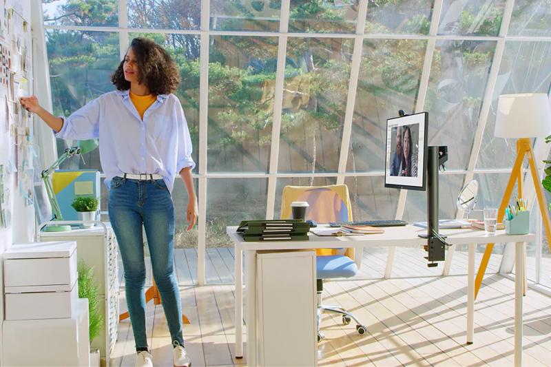 A woman in a bright office stands near a wall with papers, beside a desk with a computer and office supplies, with large windows showing greenery outside