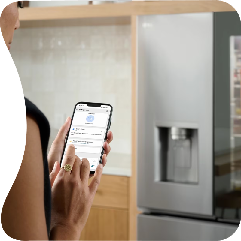 A woman stands in front of an LG refrigerator and uses the ThinQ™ app on her smartphone.