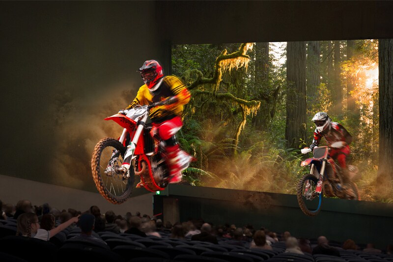 Motorcyclists appear to ride out of a movie screen in a theater, set against a forest backdrop.
