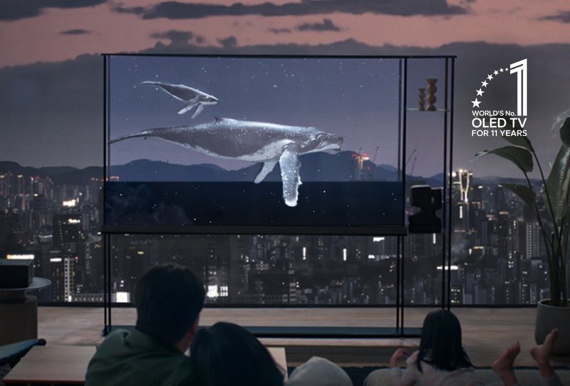 The image shows a modern living room at dusk with a city skyline in the background. A transparent OLED TV displays a scene of whales swimming underwater, making it appear as though the whales are floating in the room. The TV is sleek and minimalist, blending seamlessly into the living space. On the right side, there is a “World’s No. 1 OLED TV for 11 Years” badge, indicating the product's accolades. A family is seen watching the TV in a relaxed setting, enhancing the sense of immersion.