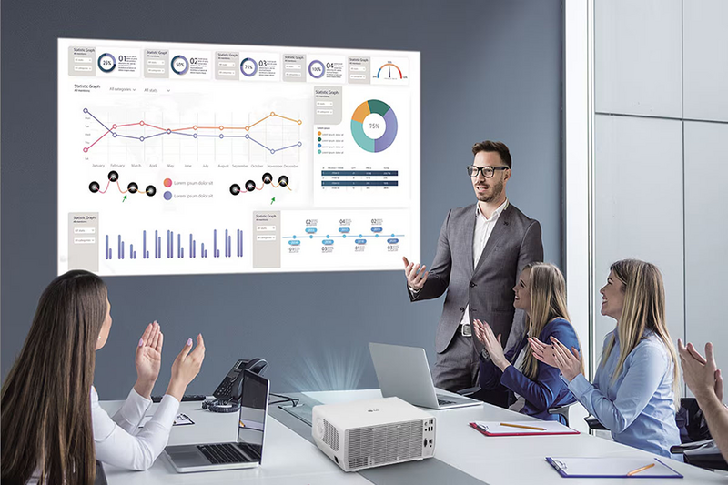 The image depicts a modern office meeting room where a group of four people is engaged in a presentation. In the center, a man in a suit is standing and gesturing towards a large projection screen displaying various charts and graphs. Three women are seated at a conference table, clapping and looking towards the presenter, suggesting that they are engaged and appreciative of the presentation. The background features a wall with a large screen displaying colorful line graphs, pie charts, and bar graphs, as well as numeric statistics. A projector is placed on the table, projecting the image onto the screen. Additional items like notebooks and pens are present, indicating an environment set for a business meeting.