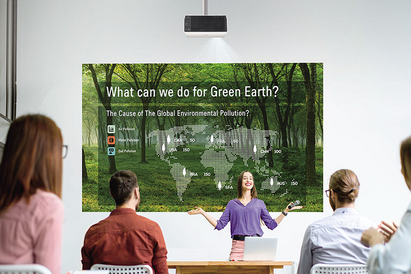 A woman presents to a group using a projector displaying a forest background with environmental data.