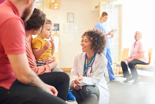 A young family sits talking to the doctor. The toddler is sat on his mother's knee as the doctor kneels down to talk to him.