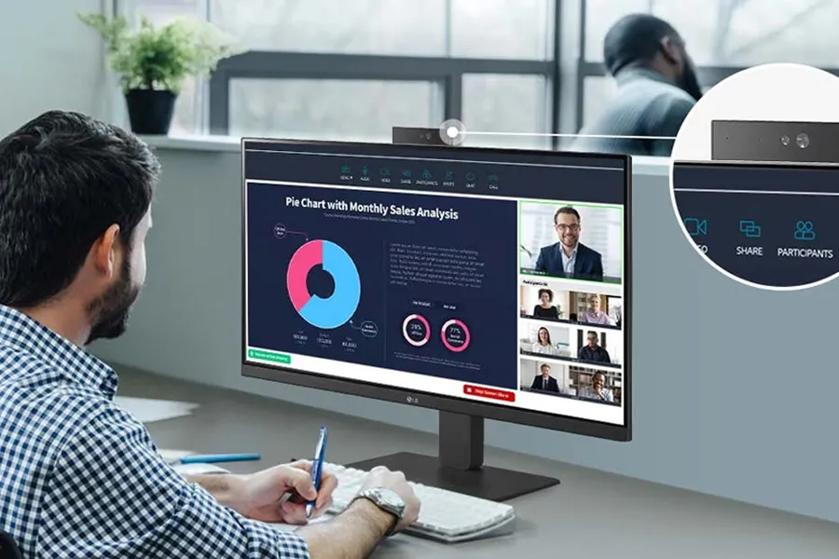 The image depicts a person seated at a desk, engaging with a large computer monitor displaying a video conference and a pie chart presentation. The background shows a window with natural light illuminating the space. The monitor screen is divided into two main sections. On the left, a large pie chart labeled "Monthly Sales Analysis" shows data in blue and pink segments. The right section features a video call interface with a main speaker's image above several smaller thumbnail images of other participants. There's a close-up inset of the webcam at the top of the monitor on the right side.