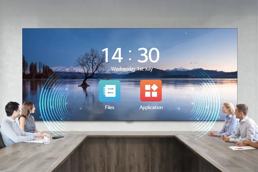 Conference room with people seated at a table facing a large digital display showing the time, date, and app icons over a lake scene.