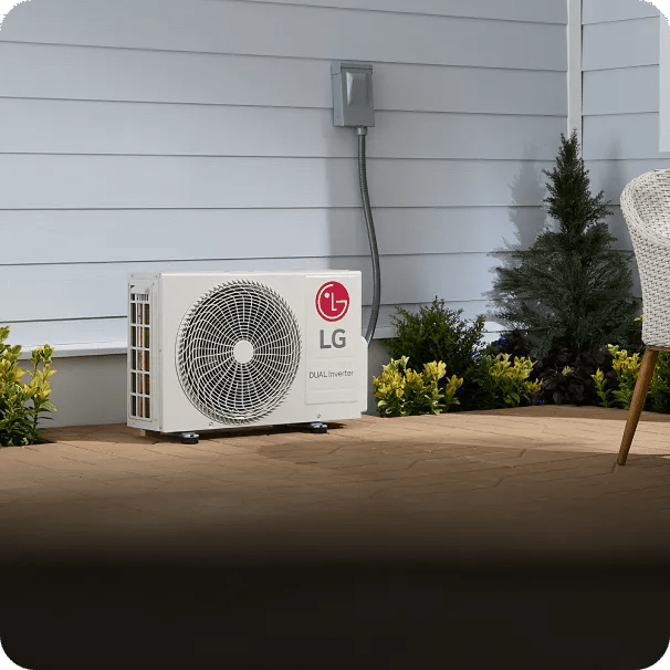 LG single-zone outdoor HVAC unit installed on a brick patio against a white-paneled house exterior, with nearby shrubs, a small evergreen tree, and part of a wicker chair visible.
