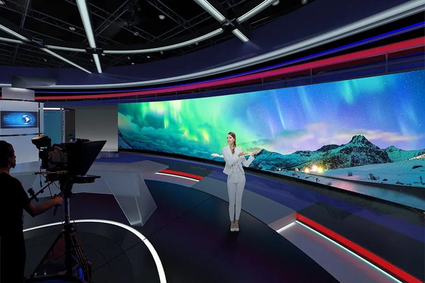 A person in a white suit stands in a modern TV studio with a large screen displaying Northern Lights.