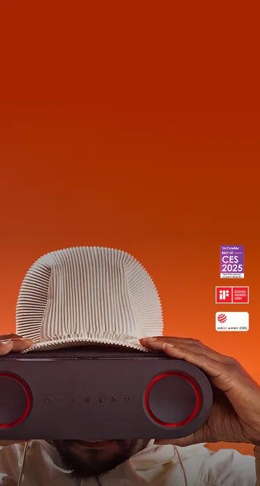 Photograph of will.i.am against an orange background faces the camera and holds an LG xboom speaker in front of his face like goggles. With three badges labeled Digital Trends 'Top Tech CES 2025,' Tech Radar 'Best of CES 2025,' and I.F. Design Award 2025.