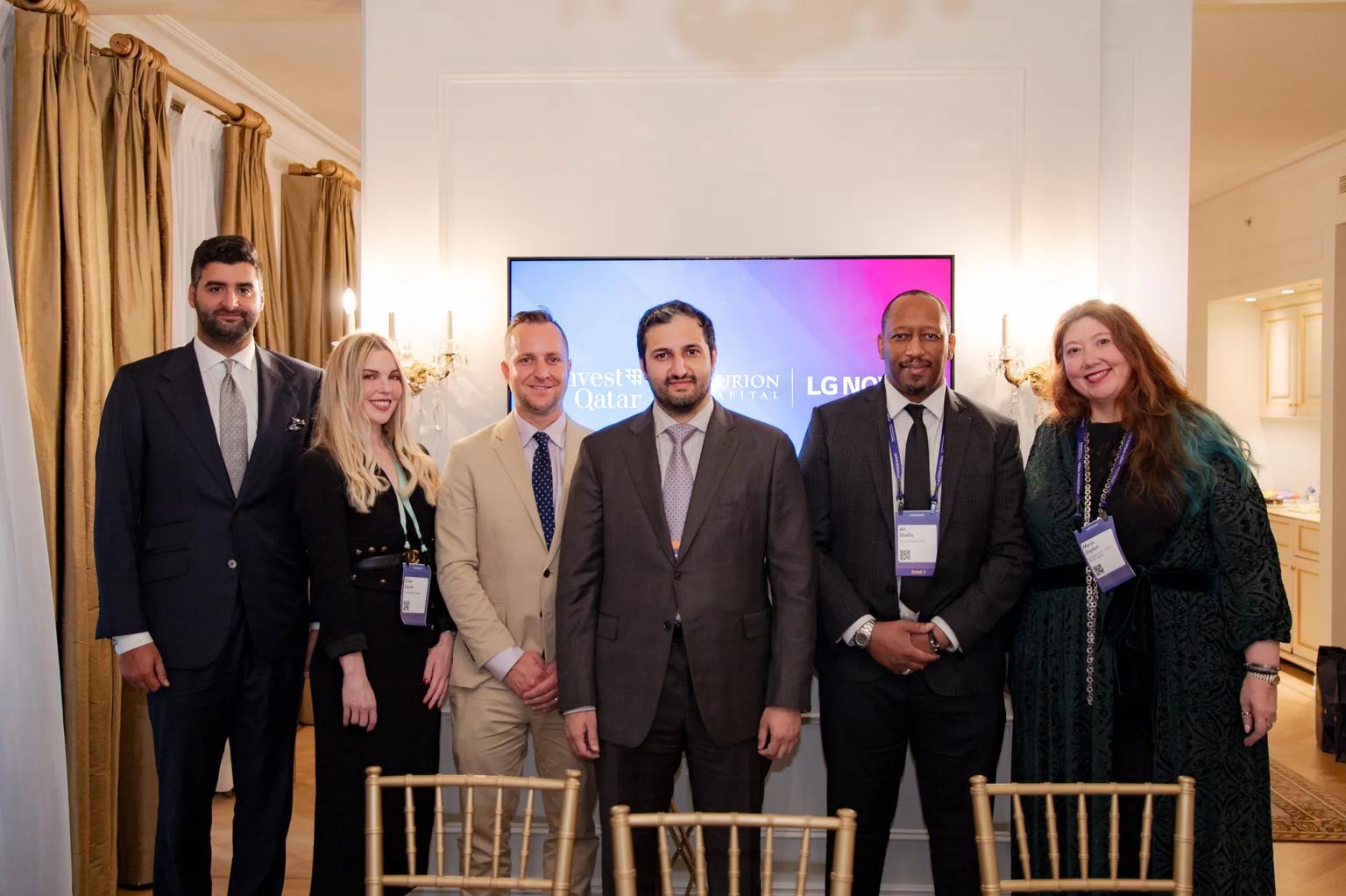 Six people pose together in formal attire for a group photo at an indoor event, standing in front of a wall-mounted screen displaying "Invest Qatar," "Falcon Capital," and "LG NOVA" logos. Several wear event lanyards with visible name badges. Gold chiavari chairs are arranged in the foreground.