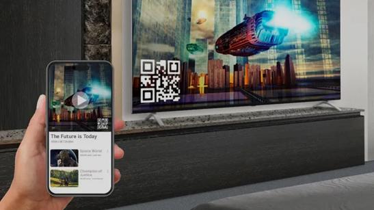 A hand holds a smart phone, showing the Apple Airplay display on it. In the background, an LG Hotel TV shows the same science fiction movie that is on the phone. A QR code on the TV facilitates the guest pairing process.