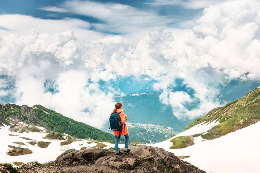 Person in an orange jacket standing on a mountain peak, overlooking clouds and distant mountains.