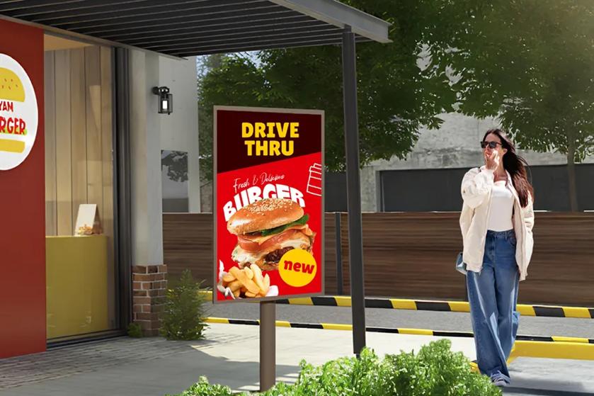 Drive-thru sign displaying a burger and fries in front of a fast-food restaurant with a woman walking by.
