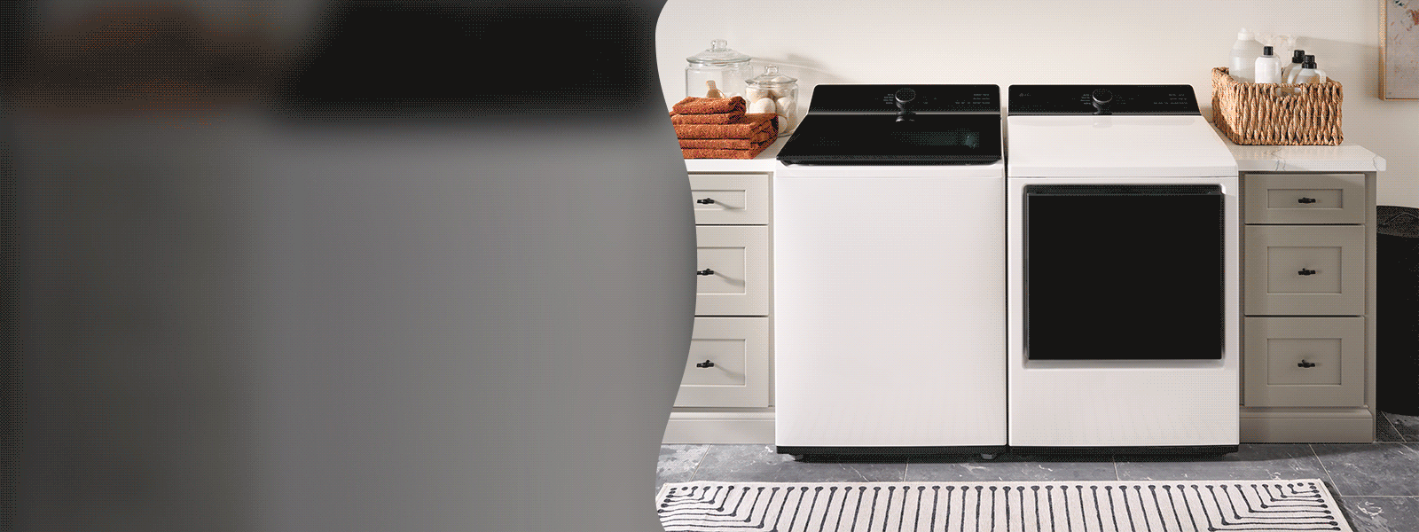 Modern laundry room with a matching LG white top-load washer and front-load dryer side by side, set between light taupe cabinets. Folded orange towels and glass jars sit on the left countertop, while a woven basket with bottles is on the right. A patterned rug lies on the dark tile floor in front.
