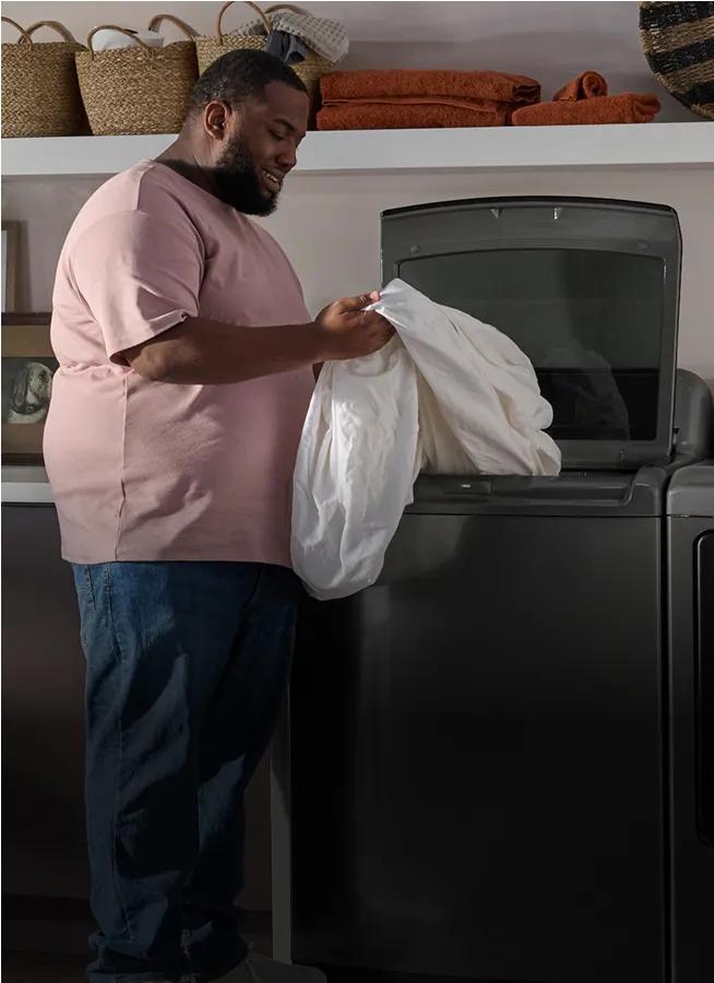 Man removing a white sheet from a dark gray top-load washer.