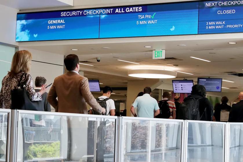 Passengers wait in line at an airport security check, while signage overhead indicates current wait times.