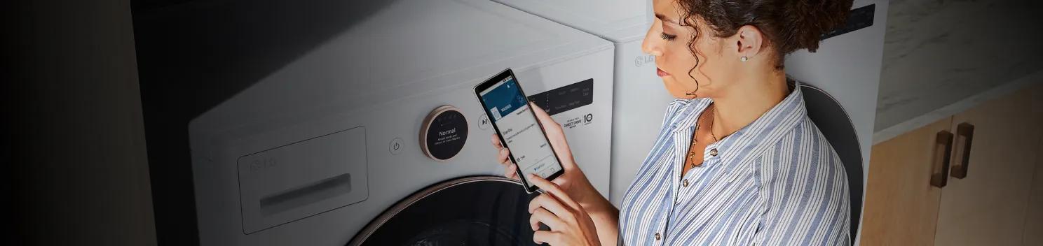 Woman standing in front of her washer and try with the ThinQ app on her phone and LG ThinQ logo at the top right.