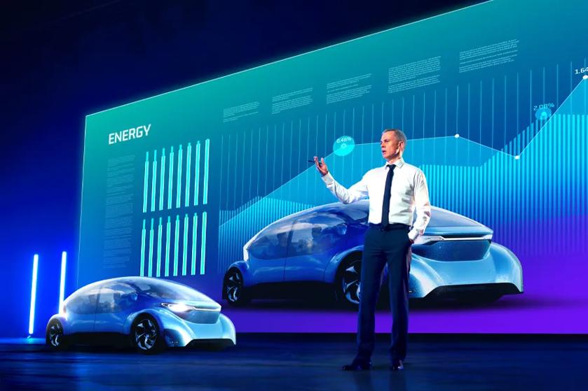 A businessman gives a presentation onstage. To his right is a sleek car, and behind him is a large scale DVLED display showcasing the vehicle features. 