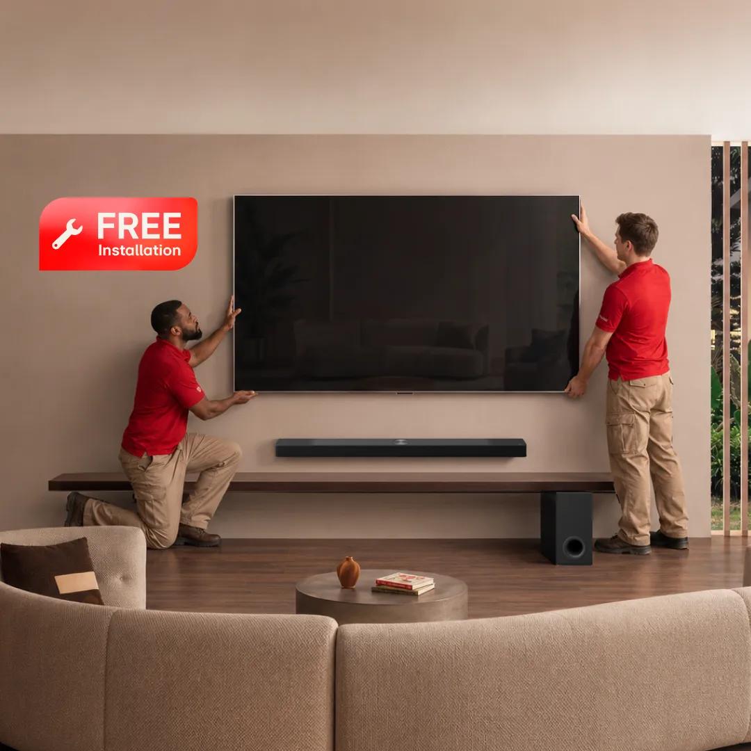 Two installers in red shirts mount a large flat-screen TV on a living room wall above a soundbar and subwoofer. “Free Installation” badge.