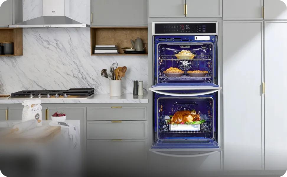 An LG double wall oven is open in a brightly lit modern kitchen. The top oven is baking pies and the bottom oven is roasting a chicken.