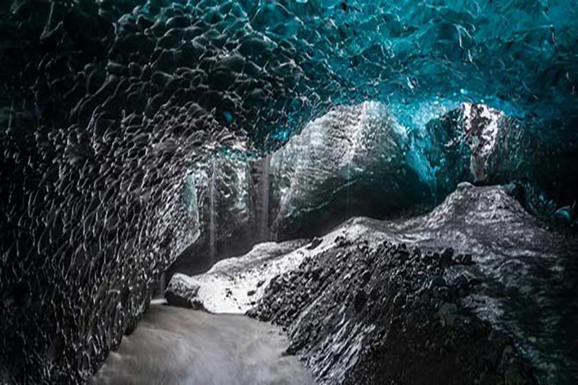 Interior of a turquoise ice cave with textured ice formations and a flowing stream.