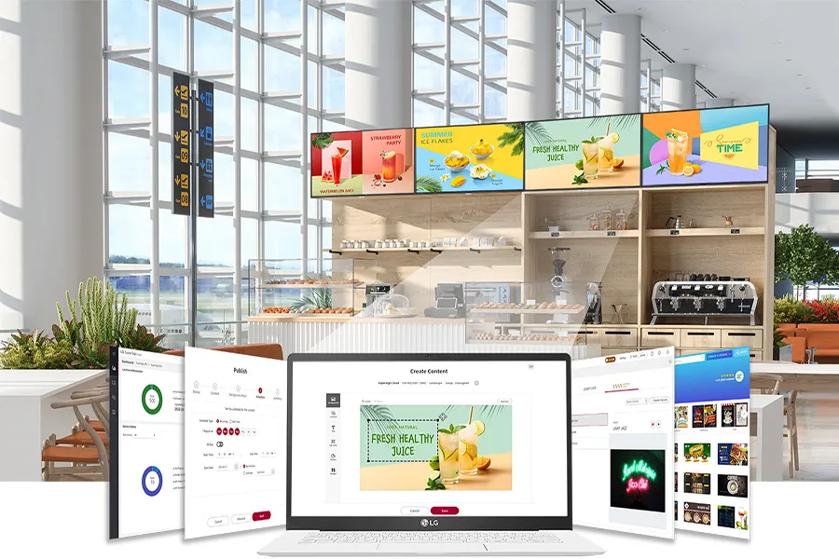 The image depicts a modern, bright café setting with large windows allowing natural light to flood the space. At the back, several digital menu boards display various colorful ads for beverages, such as strawberry drinks, healthy juice, and lemonades, each with vibrant graphics. Below the screens is a serving counter with shelves and an espresso machine. In the foreground, digital screens and a laptop are displaying software interfaces for designing and publishing content like menus and promotional materials. The space is clean and contemporary, with some green plants decorating the area.