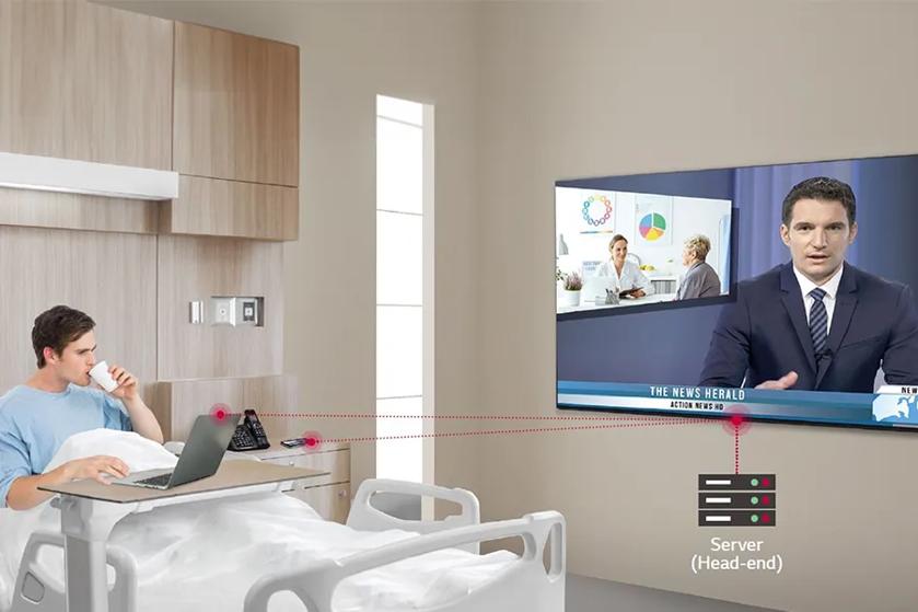 A hospital patient using a laptop, watching a news broadcast on a wall-mounted TV with network connections shown.