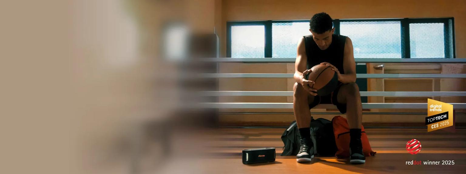 A man rests on a bench while holding a basketball with an xboom speaker nearby. “Digital Trends Top Tech CES 2025” and “reddot winner 2025” badges.