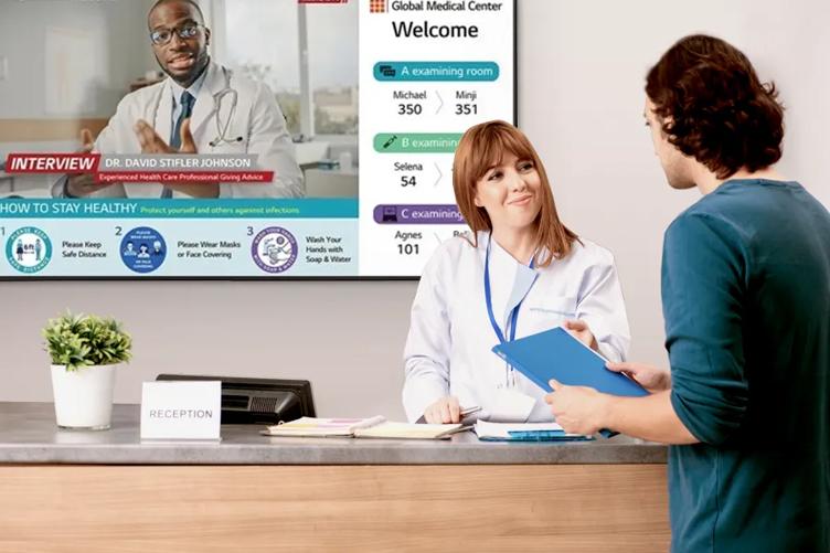 A nurse greets a patient at a hospital reception desk. An LG LCD signage is mounted on the wall behind her, displaying hospital information. 