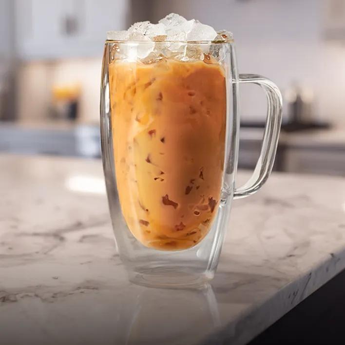 Glass mug filled with an iced coffee beverage topped with crushed ice, sitting on a marble countertop in a softly lit kitchen.