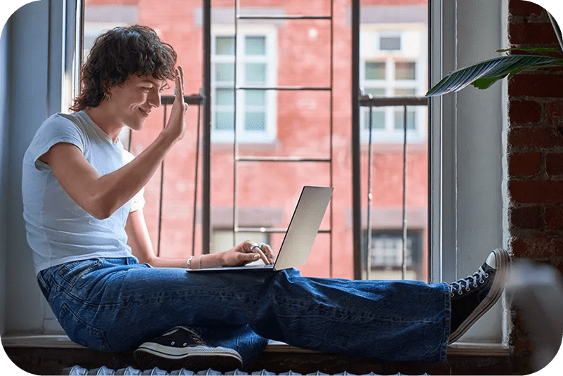 Young man sitting by an open window, smiling and waving at a LG gram as if on a video call.