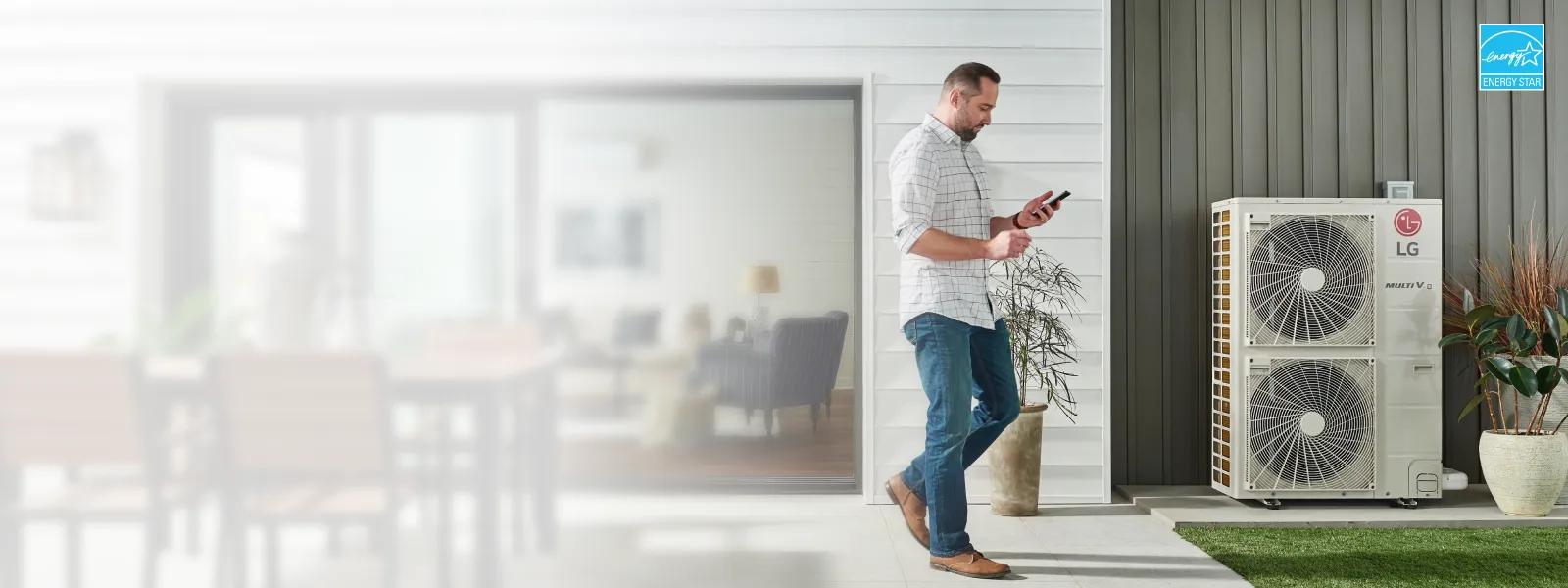 Man looking at a smartphone while standing near an LG MULTI V outdoor HVAC unit installed outside a modern home with a combination of white and gray siding, decorative potted plants, and a neatly maintained lawn. In the upper right corner, the Energy Star badge of certification.