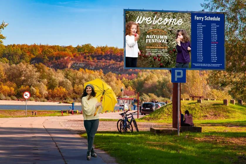 A woman with an umbrella walks past an outdoor DVLED billboard in an autumnal park scene. 