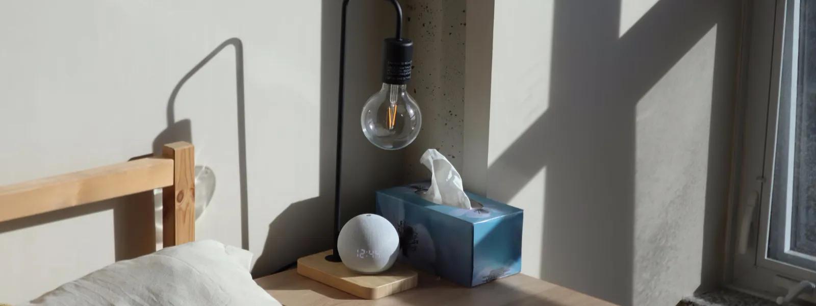 Bedroom space partially showing a bed’s headboard and pillow with a side table next to it with decorative dimmable globe bulb charging WiFi Smart lamp, and facial tissue box.