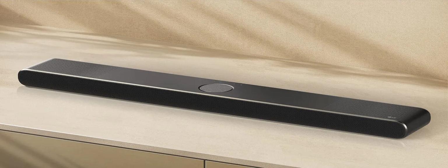 The image features a sleek black LG Soundbar with a minimalist design, rounded edges, and perforated speaker grilles on both ends, sitting on a beige surface.
