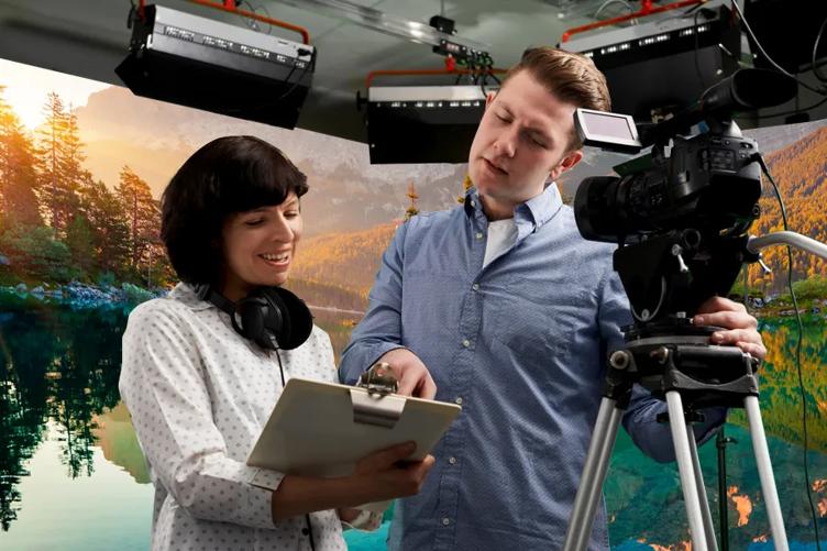 A studio manager shows a cameraman something on her clipboard. Behind them, the walls are made of LG Studio Series displays, showing a scene of a mountain lake. 