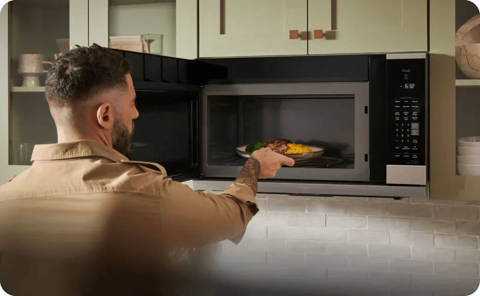 Person reaches into an open LG countertop microwave to place a plate of food inside, with the control panel and timer visible at right side.