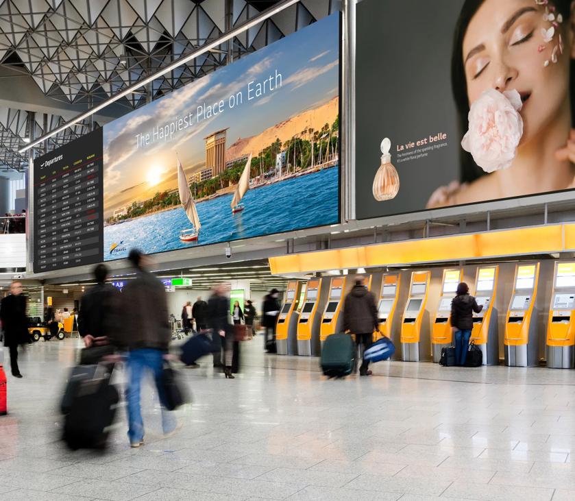 Busy airport terminal with travelers, two large ads, a departures board, and check-in kiosks.