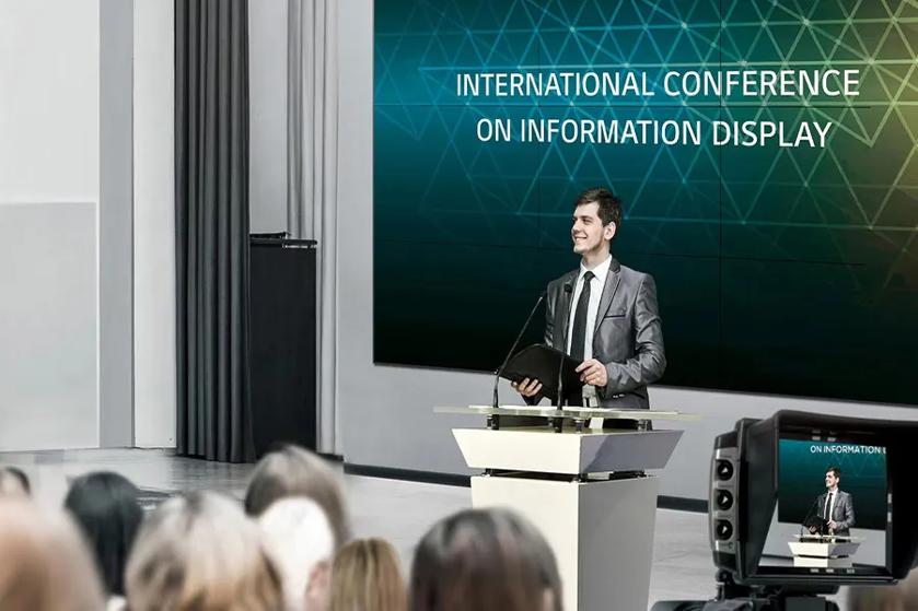 The image shows a conference setting with a speaker standing at a podium. The speaker, a man in a grey suit, is smiling and holding a black folder. He stands in front of a large screen displaying the title "INTERNATIONAL CONFERENCE ON INFORMATION DISPLAY" in white capital letters. The screen has a geometric pattern with a gradient background transitioning from blue to green. In the foreground, there is an audience, slightly blurred, indicating focus on the speaker. A video camera to the side is filming the event, with its display screen showing the speaker at the podium. The room has a modern design with grey curtains and a light color scheme.