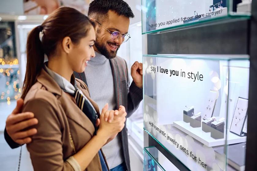 A couple smile as they look at a jewelry case. Transparent OLED over the case reads 'say I love you in style with romantic sets for her'