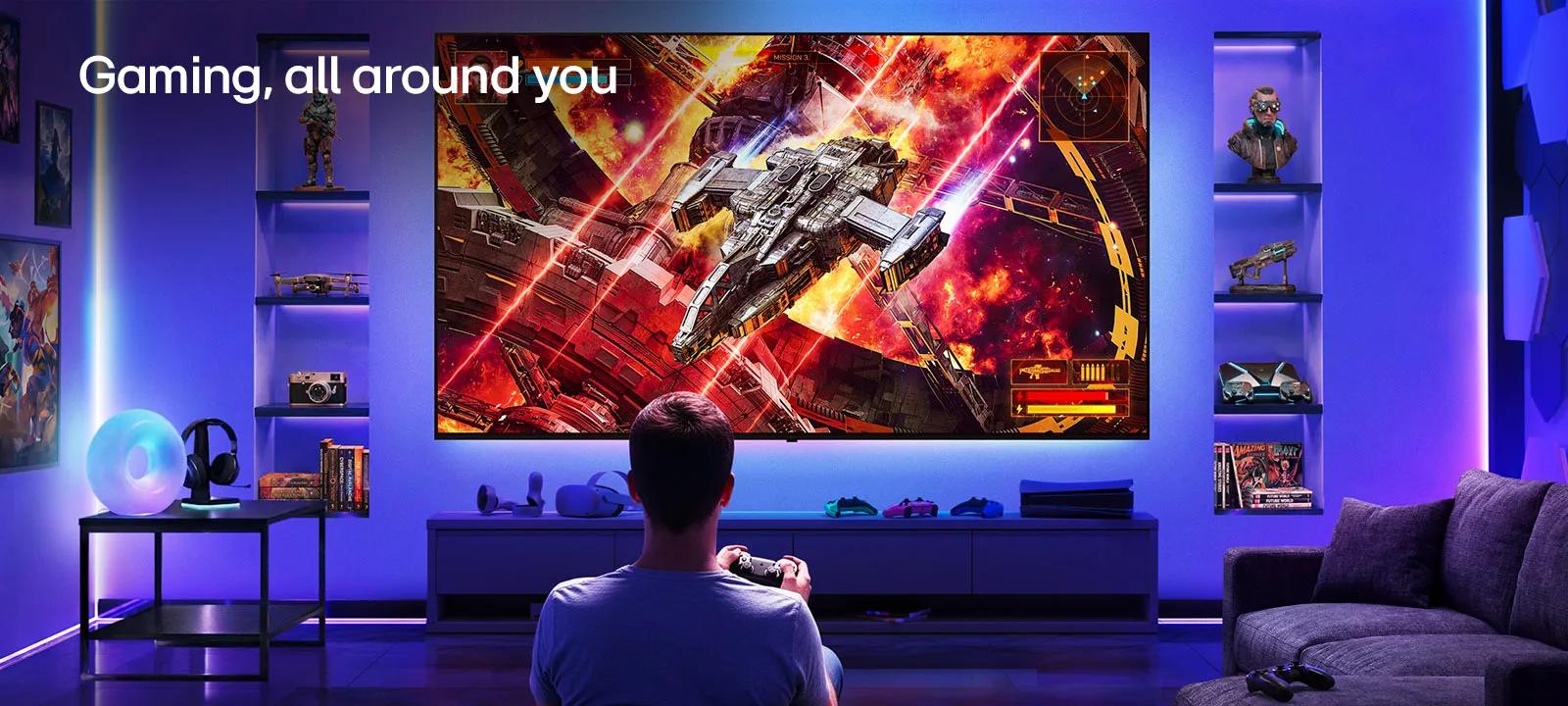 LG Ultra Big TV is wall-mounted in a neon-lit gaming room, where a large display shows a vivid space battle scene as a player sits facing the screen, illustrating an immersive and engaging gaming experience.