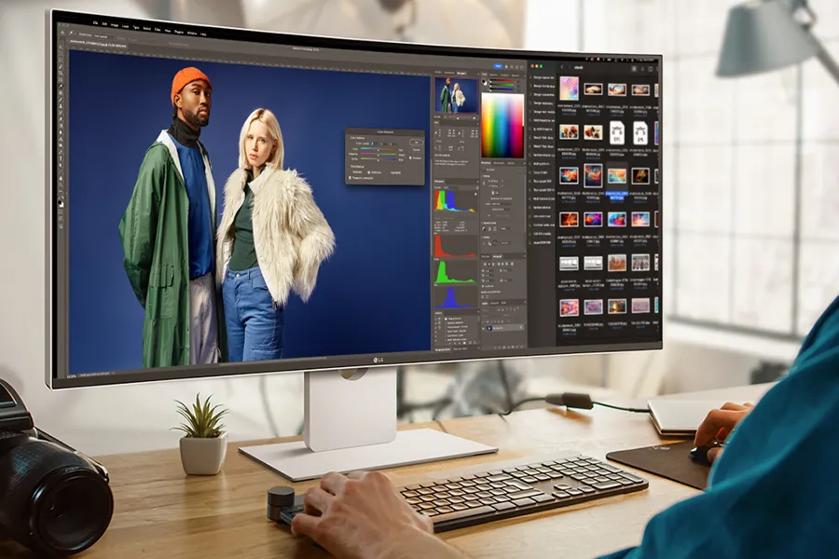 A large monitor displays photo editing software with an image of two individuals on the screen. A person is working at a desk with a plant and camera nearby.